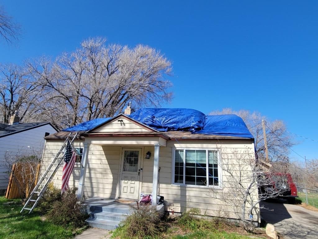 Blue emergency tarp covering the roof of a house, seen from the front, with a ladder at the eave and snow on the ground.