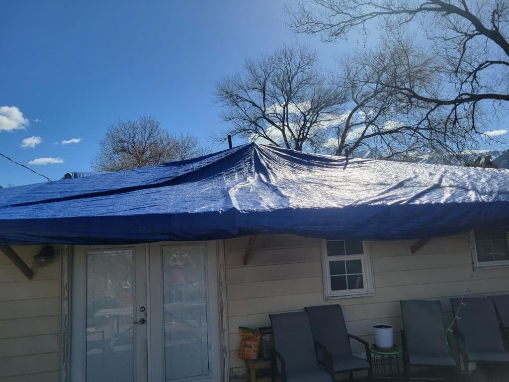 Blue emergency tarp covering the roof of a two-story house, with snow on the ground and mountains behind it.